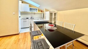 A modern kitchen with white cabinets, a black countertop, and a dining area with a black table and white chairs. - Rooftop Elegance Suite (Budva)