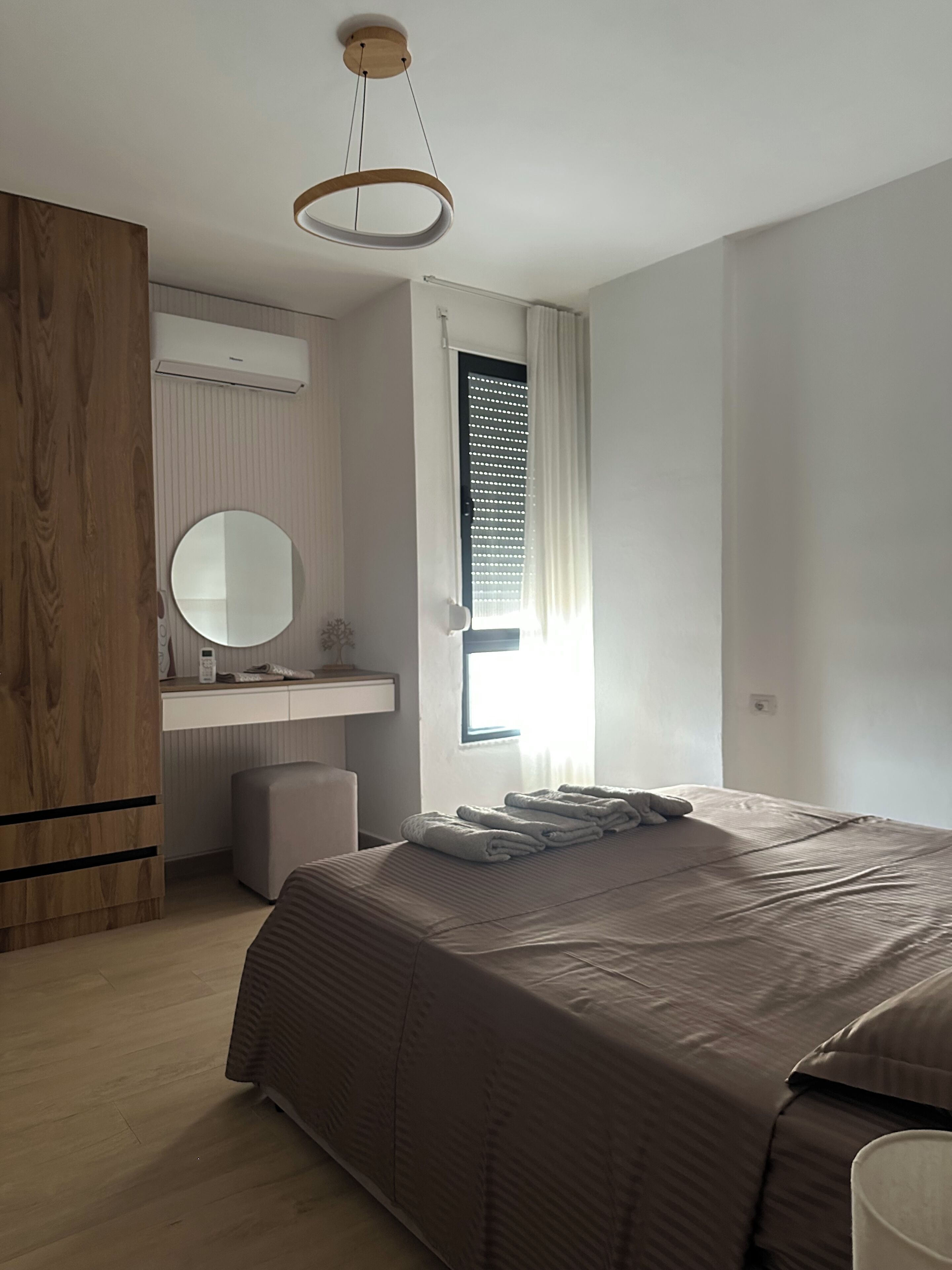 Une chambre à coucher comprenant un lit, une table de chevet, un miroir, une armoire en bois et une fenêtre avec des stores.