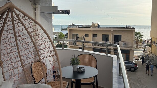 Uma varanda com uma cadeira suspensa, uma mesa pequena com uma planta e vista para o mar e edifícios.