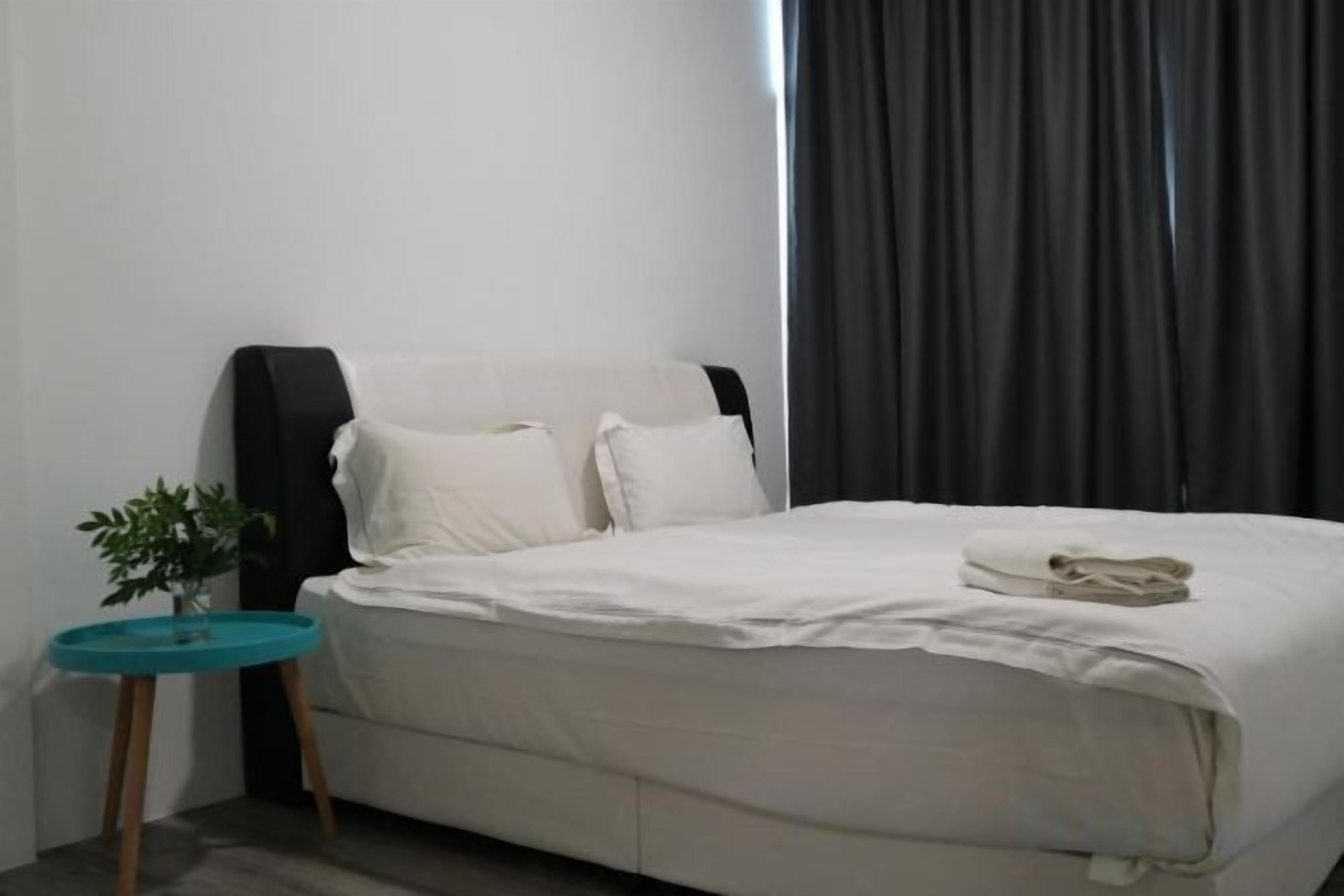 A bed with white bedding, a small round table with a plant, and a dark curtain in the background.