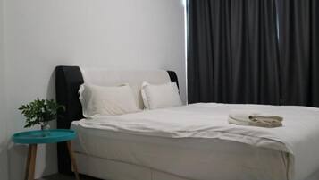 A bed with white bedding, a small round table with a plant, and a dark curtain in the background.