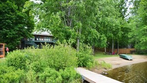 Eine Holzstege führt in einen Teich, umgeben von dichtem Grünwerk und einem Haus mit grüner Dachbegrünung im Hintergrund.
