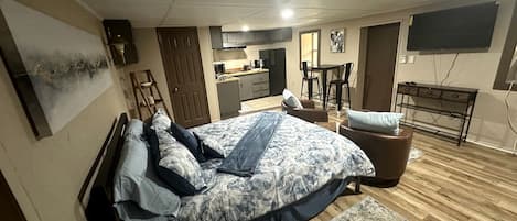 A bedroom with a bed, a desk, a chair, a TV, and a kitchen area.