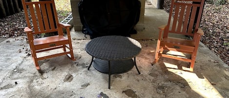 Dos sillas de madera y una mesa redonda en un patio de concreto con una parrilla al fondo.