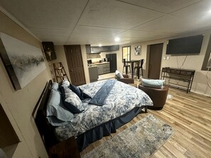 Un dormitorio con cama, escritorio, silla, televisor y una zona de cocina.