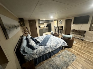 A bedroom with a bed, a desk, a chair, a TV, and a kitchen area.