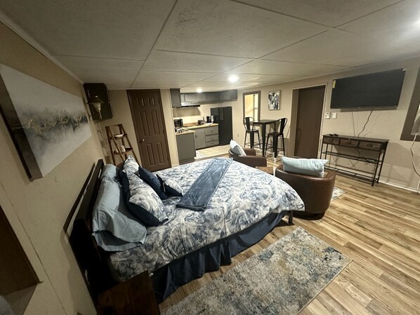 A bedroom with a bed, a desk, a chair, a TV, and a kitchen area.