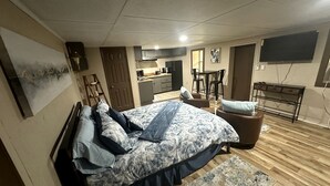 A bedroom with a bed, a desk, a chair, a TV, and a kitchen area.