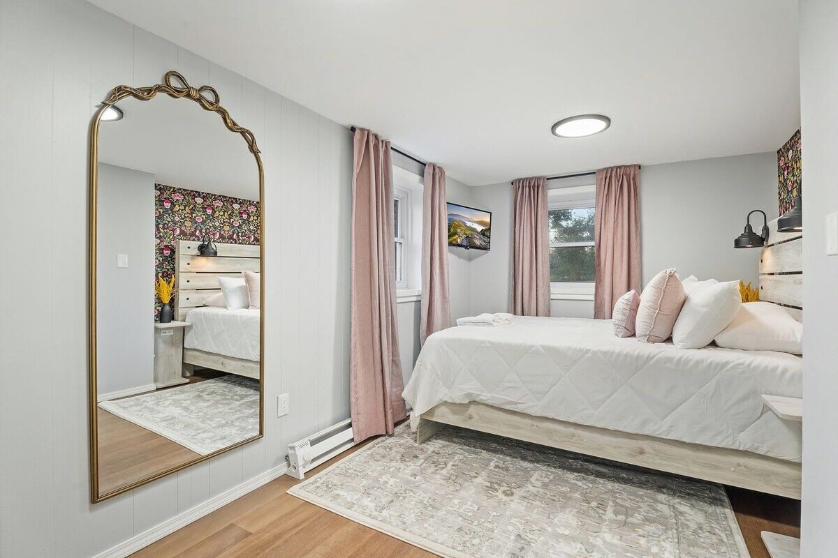 Une chambre avec un grand miroir, un lit avec une literie blanche, une fenêtre avec des rideaux et un radiateur.