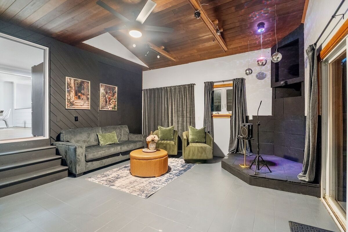 Ein modernes Wohnzimmer mit einem grauen Sofa, grünen Sesseln, einem Holztisch und einem großen Fenster mit Vorhängen.