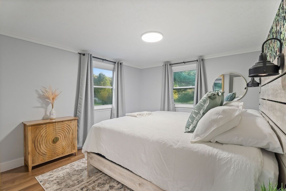 A bedroom with a bed, a wooden sideboard, a mirror, and two windows with curtains.