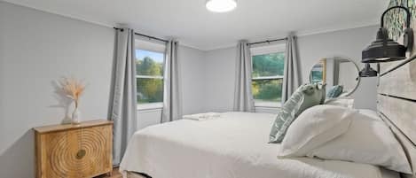 A bedroom with a bed, a wooden sideboard, a mirror, and two windows with curtains.
