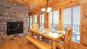 A wooden cabin interior with a stone fireplace, wooden dining table with benches, and large windows with blinds. - Bearly Chocolate (Sevierville)
