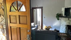 Une porte en bois avec un panneau de verre, un mur en pierre, un ventilateur de plafond, une cheminée en pierre, un canapé gris et une petite table ronde.