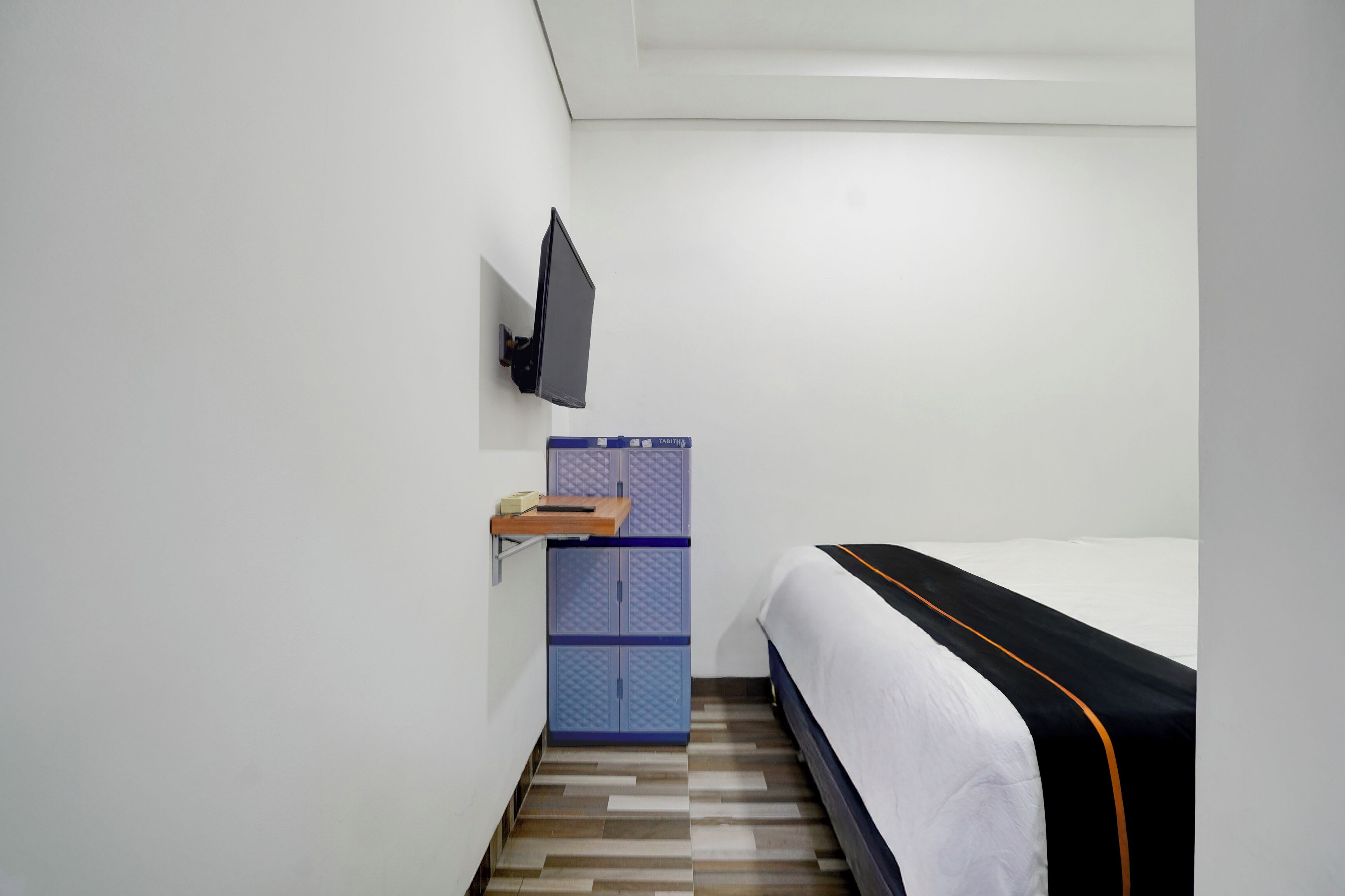A small, modern hotel room with a bed, a TV mounted on the wall, and a storage unit with a shelf.
