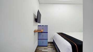 A small, modern hotel room with a bed, a TV mounted on the wall, and a storage unit with a shelf.