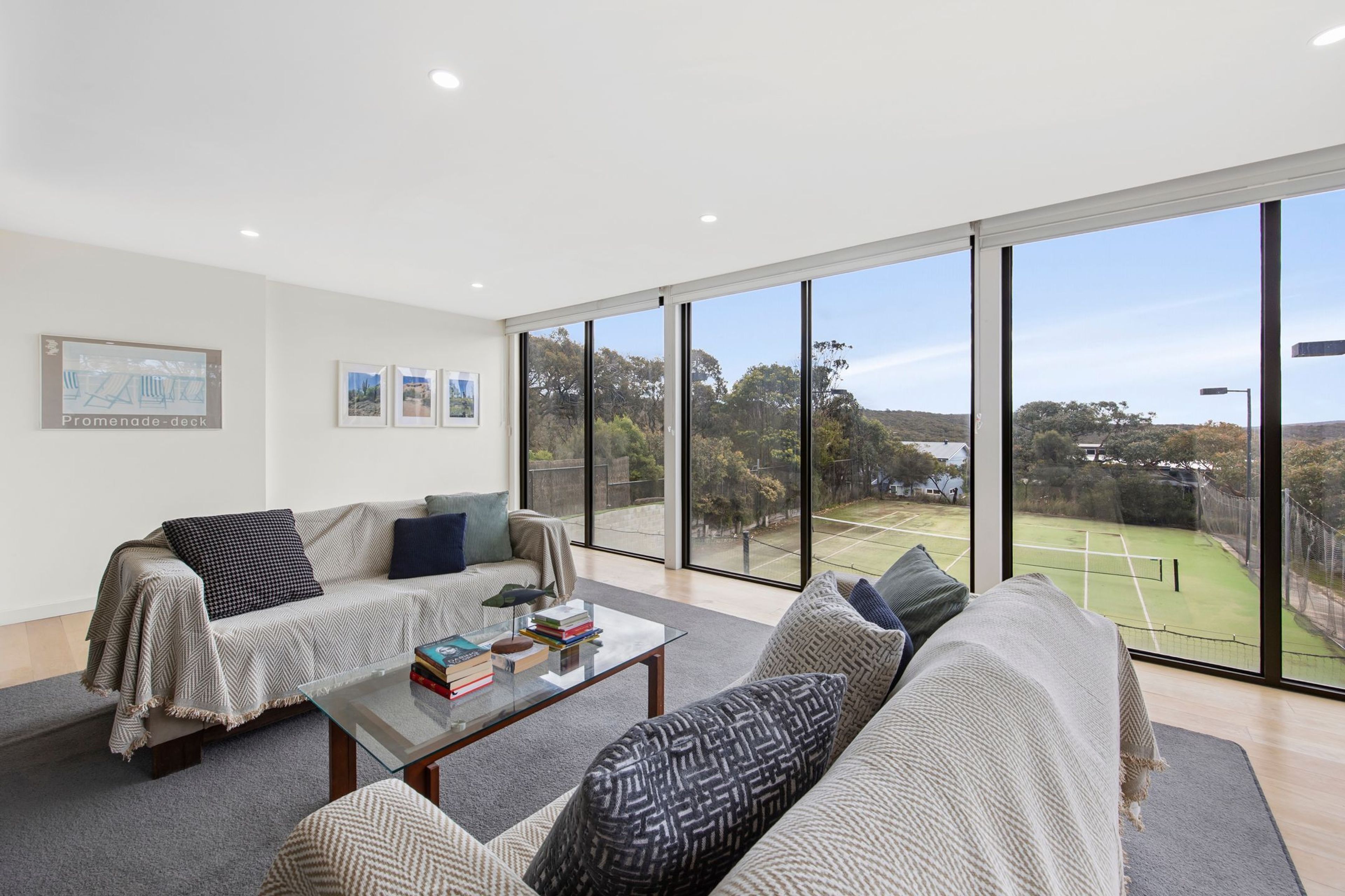A spacious living room with a large window, a sofa, a coffee table, and a tennis court view.