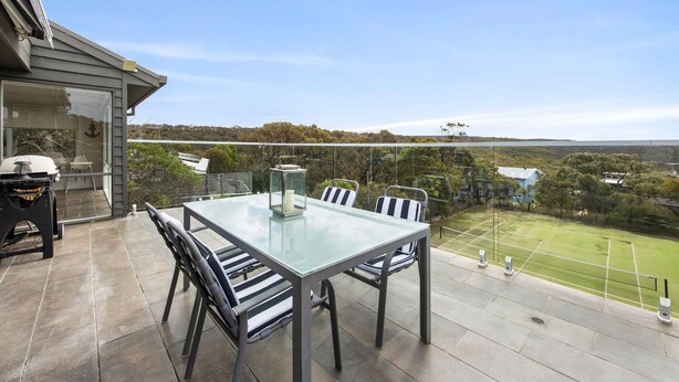 An outdoor patio with a glass table and chairs, a barbecue grill, and a view of a tennis court and distant landscape. - Rally House Fairhaven (Fairhaven)