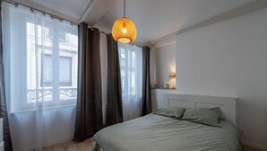 Un dormitorio con cama, cortinas, lámpara colgante y ventana con vistas a edificios.