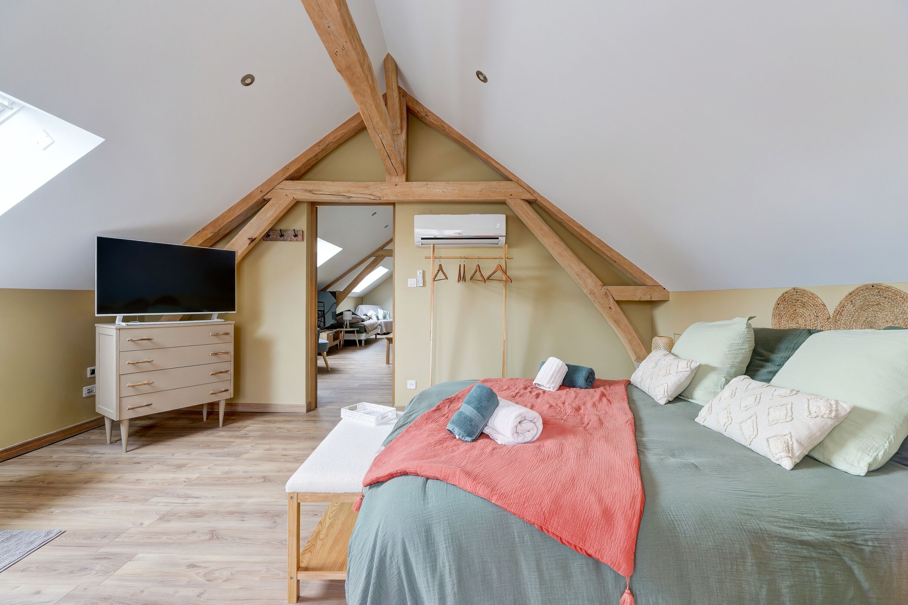 Un dormitorio con techo inclinado, televisor de pantalla plana, cómoda de madera, cama con almohadas y manta, y equipo de aire acondicionado.