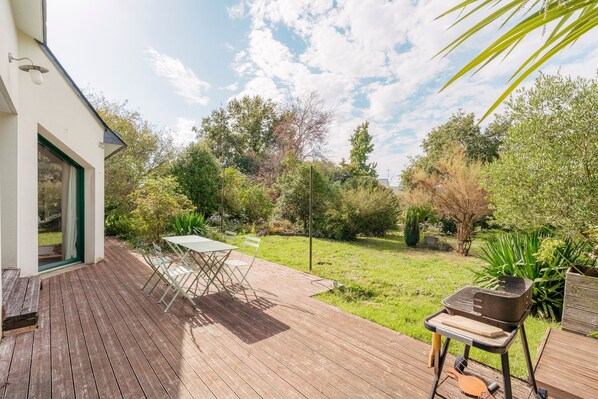 Eine Holzterrasse mit Tisch und Stühlen, einem Grill und Blick auf einen Garten mit verschiedenen Pflanzen und Bäumen.