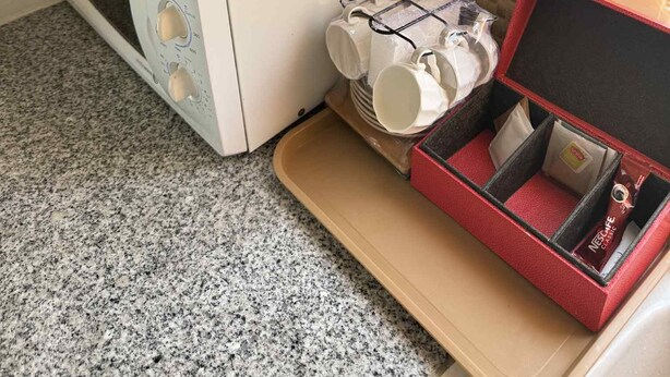 Et tekøkken med mikrobølgeovn, kaffemaskine og en kaffemachine på en bordplade. Der er en bakke med kopper og en æske med en kaffekapsel.