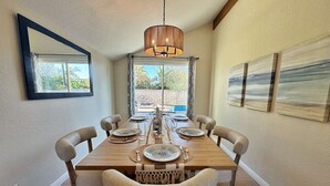 Uma sala de jantar com uma mesa de madeira posta para seis pessoas, seis cadeiras acolchoadas, um candelabro pendente e vista para o exterior pela janela.