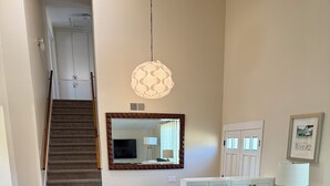 Uma sala de estar com um sofá azul escuro, duas cadeiras de madeira, uma mesa de centro redonda de vime, um espelho grande e uma escada ao fundo.