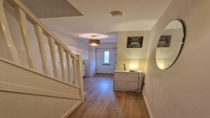 Un couloir doté d’un plancher en bois, d’un escalier blanc, d’une table de chevet avec une lampe et d’un miroir fixé au mur.