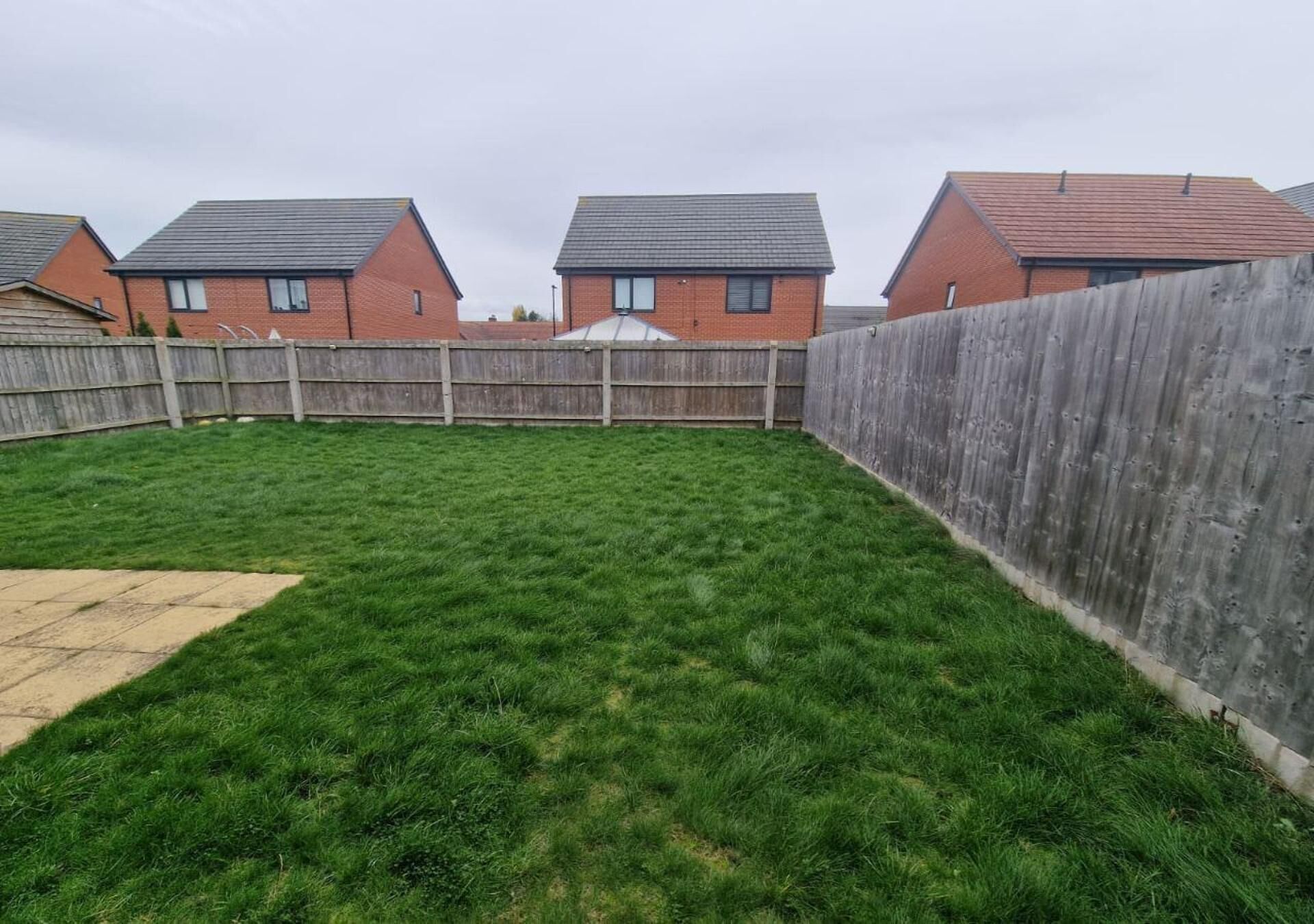A backyard with a wooden fence, green grass, and a brick house with a tiled roof.