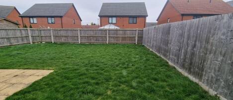 A backyard with a wooden fence, green grass, and a brick house with a tiled roof.