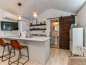 A modern kitchen with a central island, bar stools, a TV, and a wooden sliding door leading to a bathroom. - Historical Tiny Home with Patio (Oklahoma City)