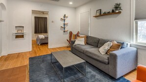 A modern living room with a grey sofa, a coffee table, and a ceiling fan. There is a window with blinds and a shelf with decorative items. - Updated Garage Apartment (Oklahoma City)