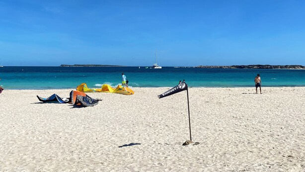 Una playa con cielo despejado, una bandera y un velero a lo lejos.