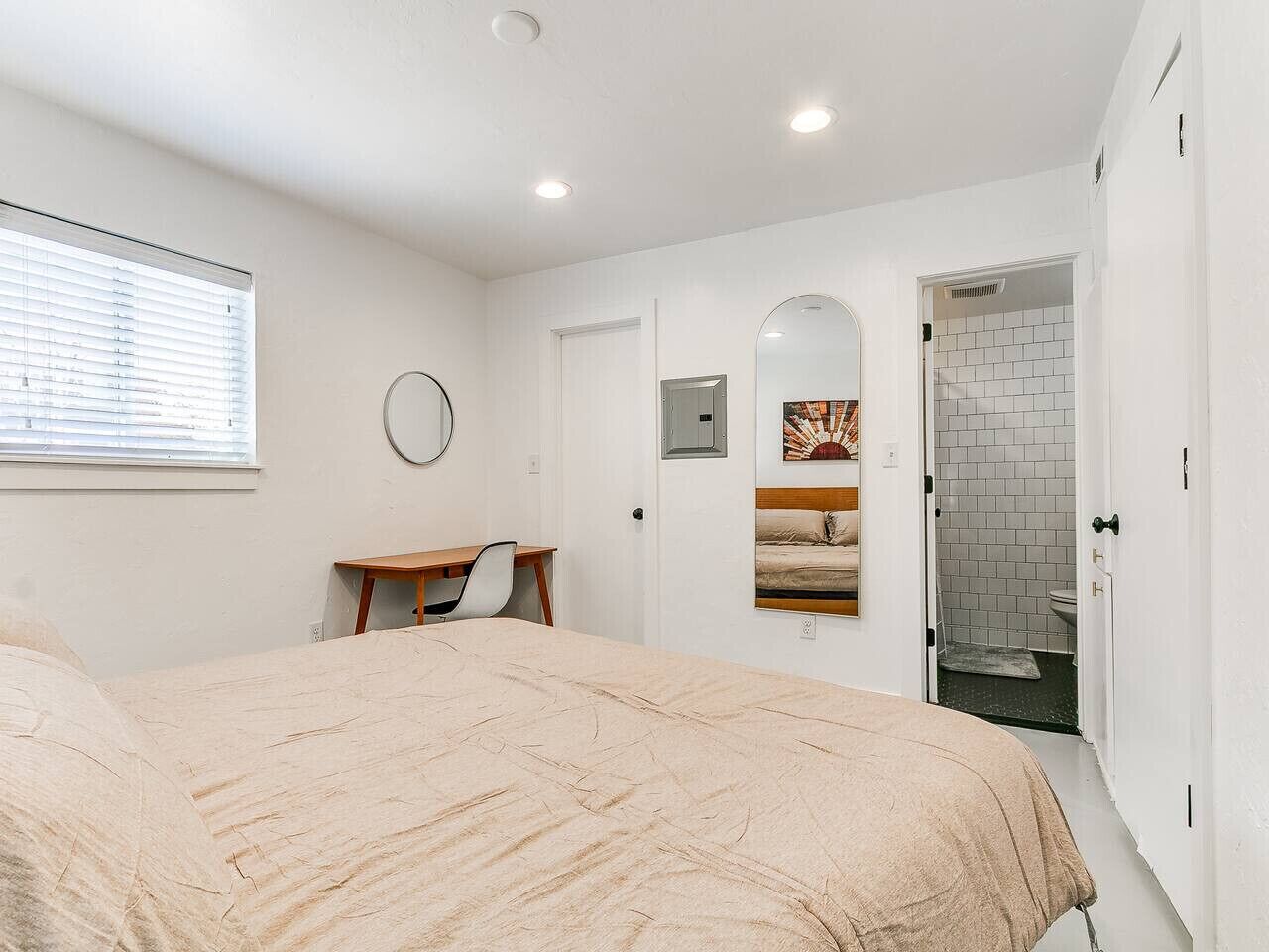 Un dormitorio con cama, escritorio, espejo y baño visible a través de una puerta abierta.
