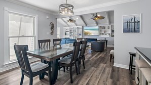 Un espace repas moderne avec une table en verre et des chaises sombres, un ventilateur de plafond et une vue sur un salon équipé d’un téléviseur et de sièges.