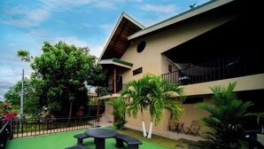 A courtyard with a green artificial turf, a stone table, and benches. There are palm trees and a building with a balcony. - Blu House Luxe: 6BR for Large Families, Jacuzzi and Panoramic Views (Provincia de Alajuela)