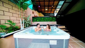 Um casal em uma banheira de hidromassagem, com parede de tijolos e plantas verdes ao fundo.