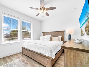 A bedroom with a wooden bed, white bedding, a ceiling fan, a nightstand with a lamp, and a beach-themed wall art. - Salty Blue (Panama City Beach)