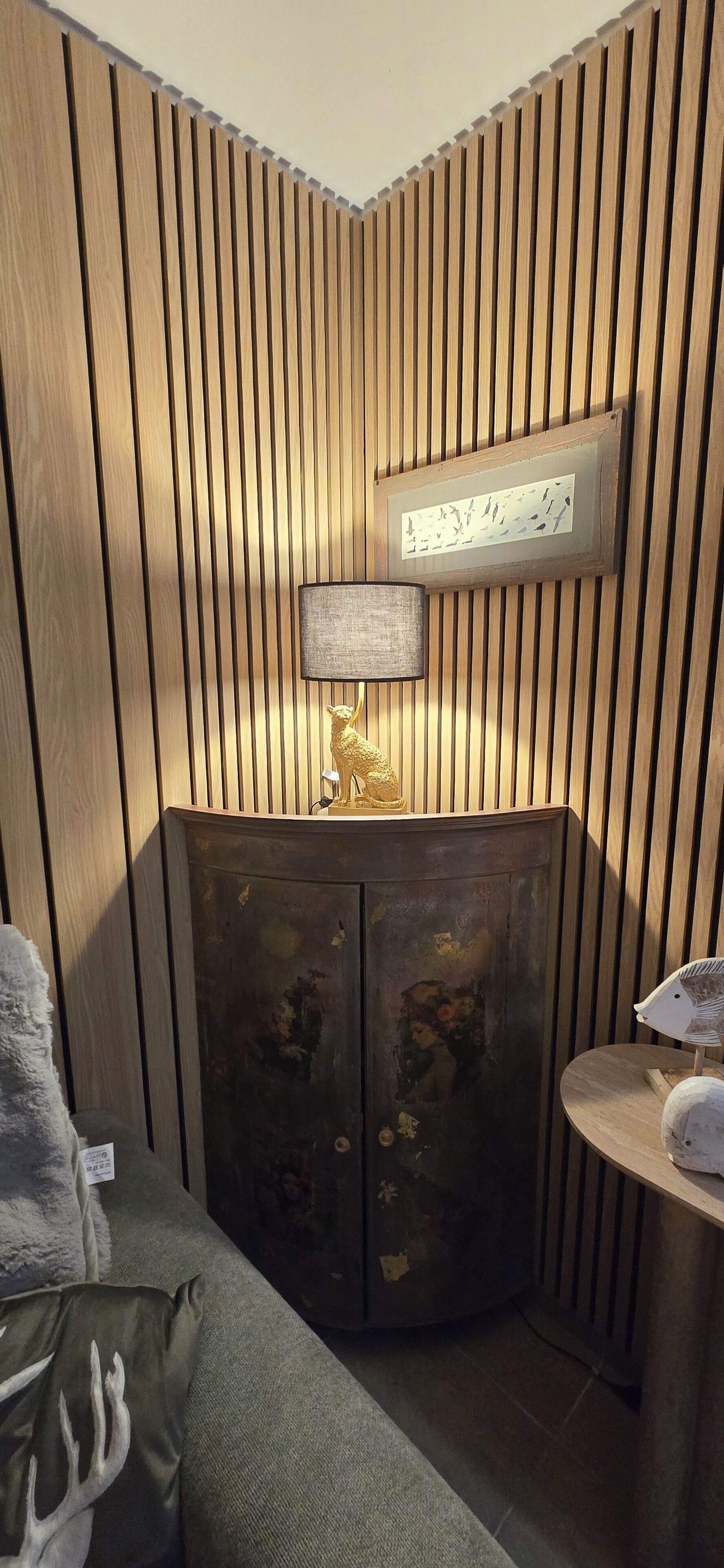 A corner room with a wooden slat wall, a cabinet with a lamp, and a small round table with a decorative item.