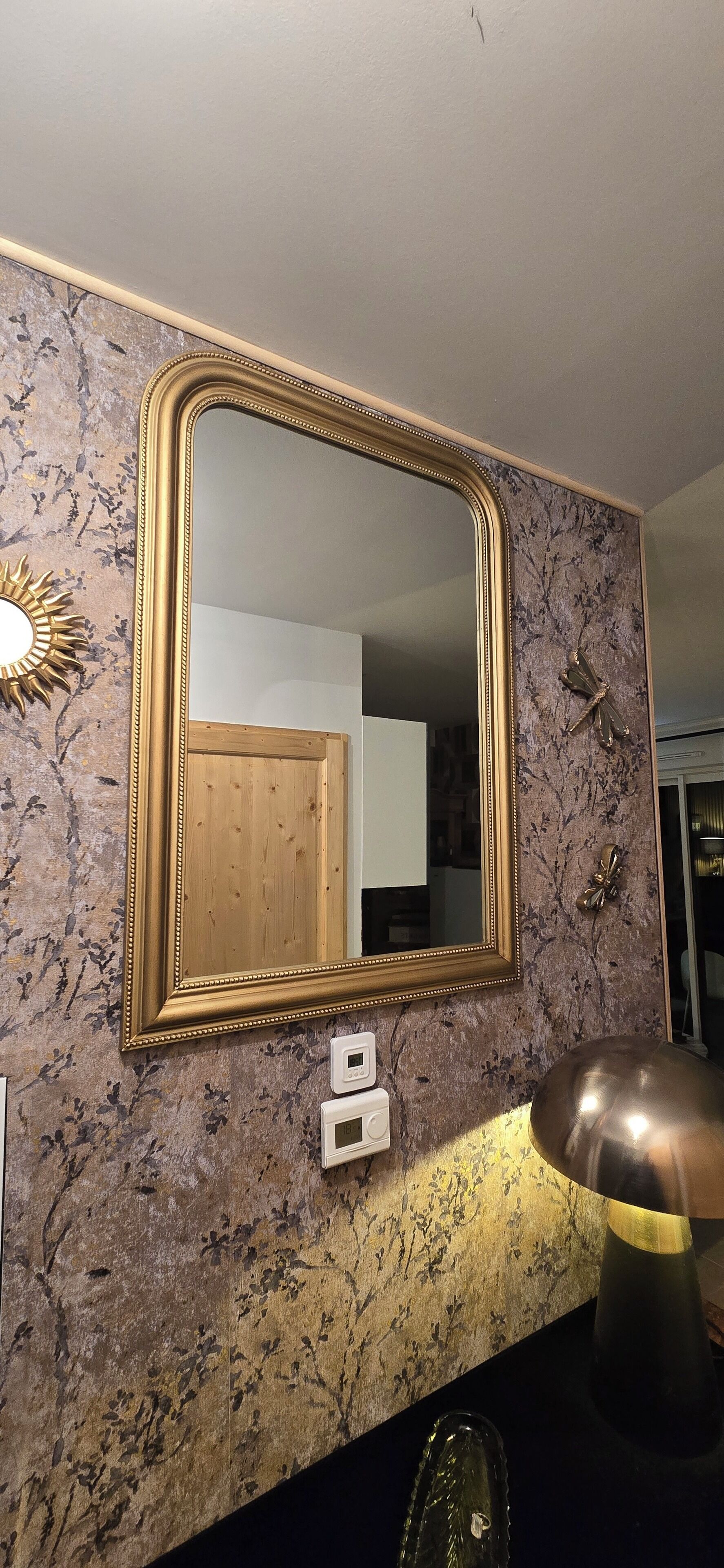 Ein Zimmer mit einem großen, goldfarbenen Spiegel, einem Thermostat und einem dekorativen Wandspiegel in Form einer Sonne.