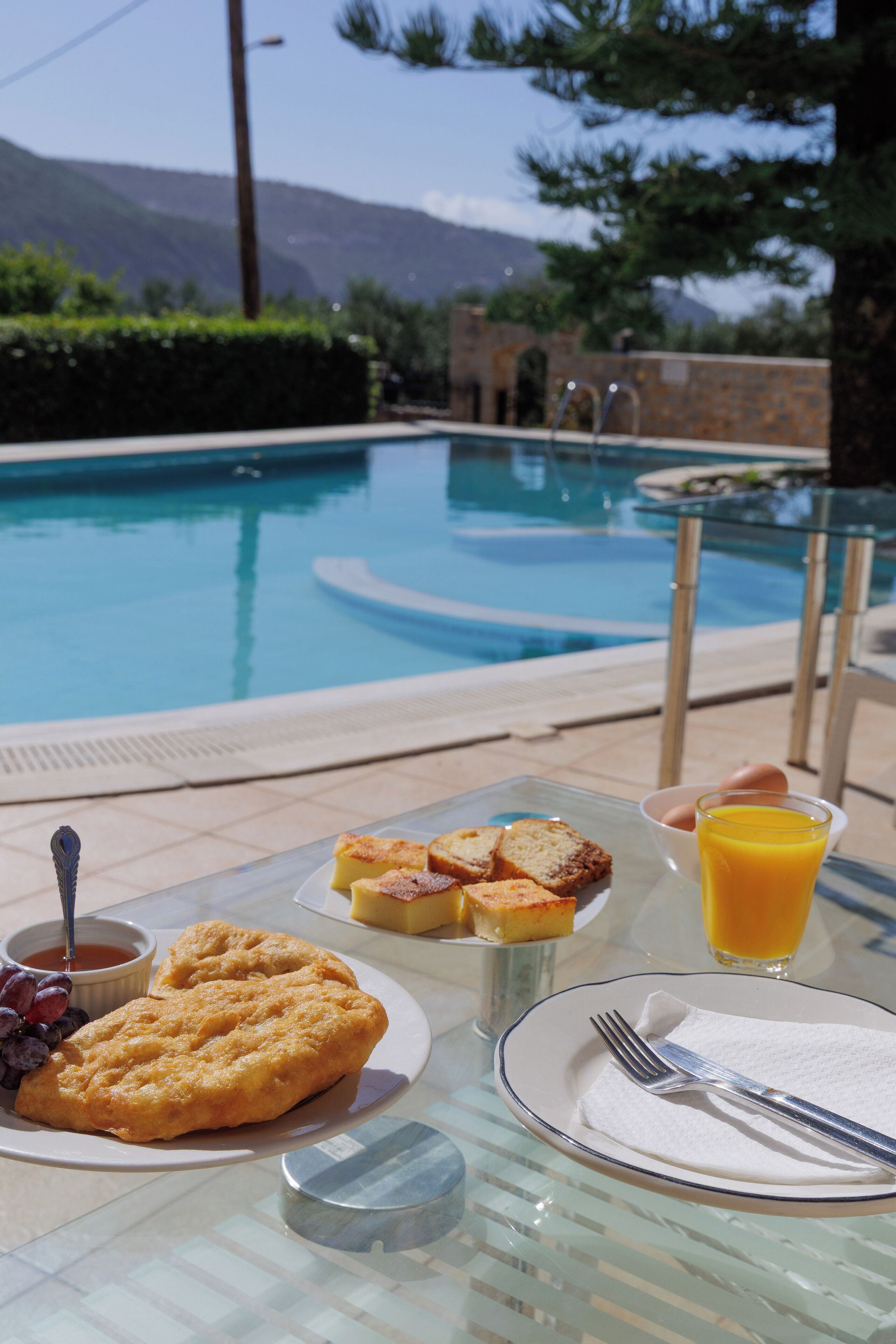A breakfast table with toast, fruit, and juice by a pool with mountains in the background.