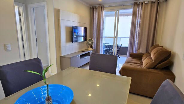 A modern living room with a sofa, a TV, a dining table with a blue centerpiece, and a balcony with curtains. - 3 minutes from the beach in the center of Bombinhas. Fully equipped apartment. (Bombinhas)