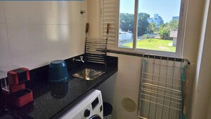 A compact kitchen with a sink, a washing machine, and a window with a view of a residential area. - 3 minutes from the beach in the center of Bombinhas. Fully equipped apartment. (Bombinhas)