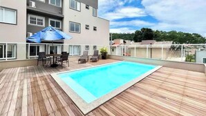 A rooftop pool area with a wooden deck, a table with chairs, and a blue umbrella. - 3 minutes from the beach in the center of Bombinhas. Fully equipped apartment. (Bombinhas)