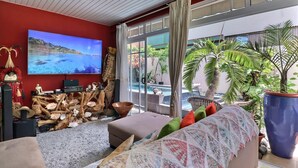 A modern living room with a large TV, comfortable seating, a view of the outdoors, and a tropical plant. - Hiiaka room - city center - swimming pool (Papeete)