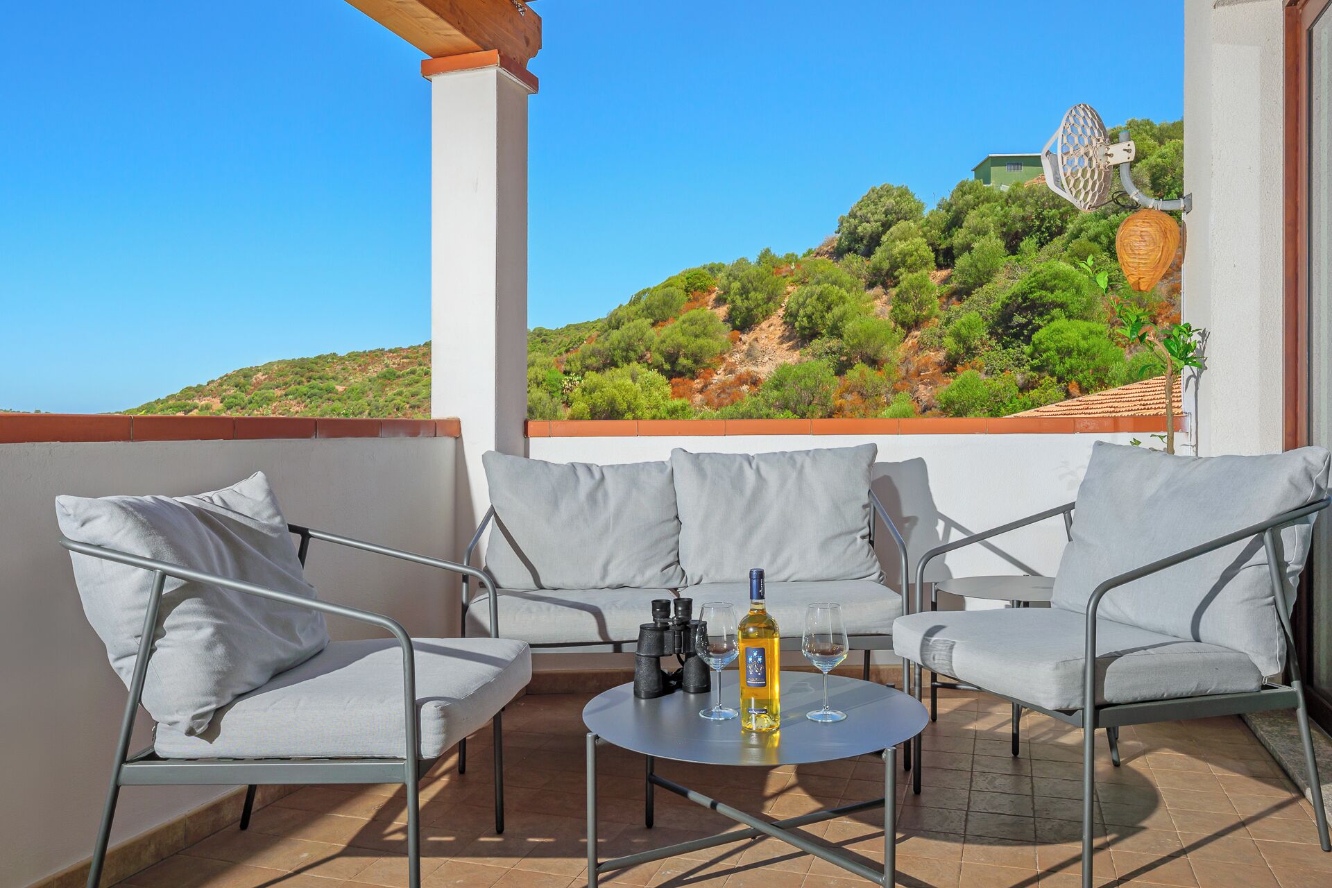 A balcony with outdoor seating, a table with a bottle and glasses, binoculars, and a view of a hillside with trees and a house.