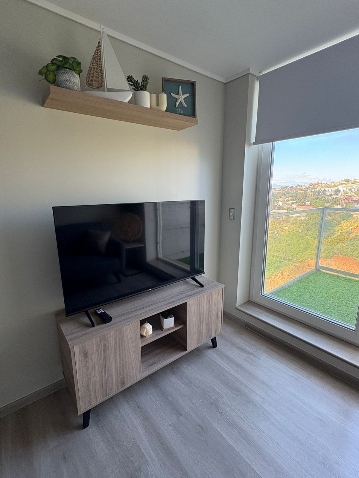 A modern living room with a flat-screen TV on a wooden TV stand, a small shelf with decorative items, and a large window offering a view of a cityscape.
