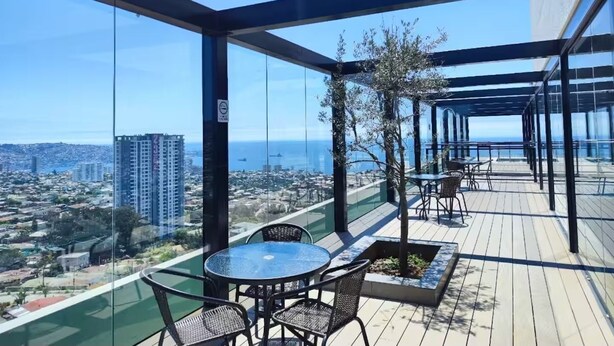 Ein Hochhaus mit einer glasüberdachten Außenterrasse, ausgestattet mit Tisch und Stühlen, das Blick auf Stadt und Meer bietet.