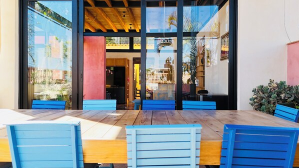 An outdoor seating area with wooden tables and blue chairs, large glass windows, and a view of the interior. - El Diablo y La Luna B&B (Oaxaca)